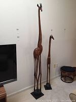 Two wooden giraffe statues standing on the floor near a wall and furniture, showing comparative height and style.