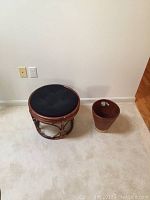 Image showing a round wooden stool with black cushion and a brown ceramic planter with sticker residue inside placed on a light carpeted floor against an off-white wall.