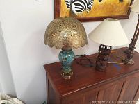 Two decorative table lamps shown on wooden sideboard; left lamp has perforated metal scalloped shade and floral ceramic base, right lamp has metal banded base and white fabric shade.