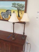 Two brass colored decorative table lamps with fabric shades, shown on wooden cabinet beneath a framed painting. Lamps show slight wear and discoloration on shades.