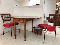 Wood gate leg drop leaf dinette table with small drawer and two metal and wicker chairs with red seat pads, shown in a home interior setting.