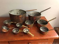 Full set of eight copper pots and measuring cups arranged on a wooden surface showing tarnish and wear.