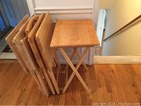 Five wooden TV tables with folding stands stacked upright next to a wall on wooden flooring.