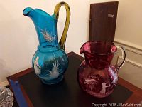 Two vintage glass pitchers; one red and one blue, placed on a black surface, showing painted white figures of girls and nature.