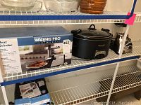 Shelf with Waring Pro meat grinder box and black Crockpot slow cooker placed next to each other