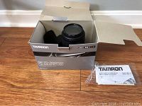 Tamron telephoto lens inside original box with user manual placed beside box on wooden floor.