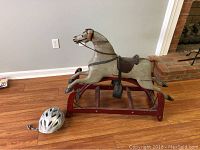 Wooden rocking horse with red base and silver Bell bike helmet on floor against gray wall.