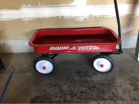 Side view of the Radio Flyer wagon, showing red metal body, white wheels with red centers, and black pull handle.