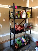 Front angle showing black metal shelving unit holding gardening and household items, with visible dents and bends on front legs and bolted construction.