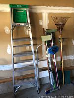 Full view of aluminum step ladder, leaf rake, push broom, round broom, traditional broom, mop, and snow shovel against garage wall.