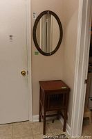 Wall mounted oval mirror with plastic decorative frame and wood accent table below in a corner.