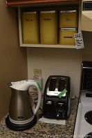 Shelf with three vintage yellow-beige metal canisters labeled Flour, Sugar, Tea, next to a T-Fal black 2-slice toaster and Oster electric kettle on countertop.