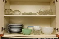 View of assorted stacked dinner plates, jadeite bowl, milk glass bowls, and glass serving pieces in cabinet shelf