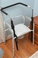 Full front view of the white metal framed toilet commode chair with padded black armrests and removable seat cover.