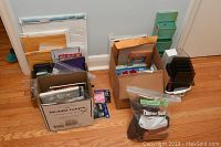Four cardboard boxes containing assorted office supplies including manila envelopes, notebooks, pens, pencils, calculators, organizers, eyeglass cases, and binders on a wood floor.
