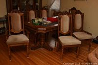 Six oak wood dining chairs around a wooden dining table on hardwood floor. Chairs have beige velour upholstered seats and backs with two captain chairs having carved wood topped backs.