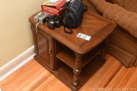Photo showing one oak wood end table next to a couch with a side cupboard door and lower shelf, with cameras and camera bag on top.