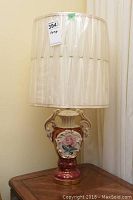 Full view of ceramic base table lamp with protective plastic cover on cream fabric shade, showing floral design on base.