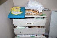 Blue-top dresser with folded assorted linens including white embroidered cloths, crocheted doilies with yellow borders, linens with brown patterned trim, and multiple folded cloths inside open drawers.