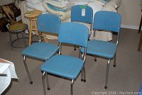 Four blue vinyl chairs with curved chrome metal legs and frames arranged in a semicircle on carpeted floor, with stools and furniture in background.