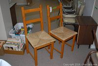 Two wooden ladder-back chairs with woven loom seats placed side by side, showing full view of chairs and surroundings including nearby boxes and other furniture.