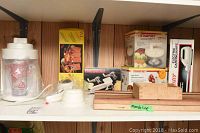 Shelf showing a white Oster juicer with plastic measuring cup wrapped, wooden mandoline slicer labeled 'mandoline', electric carving knife box, and other kitchen items.