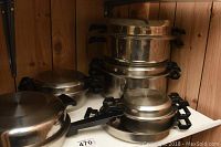 Six stainless steel Life Ware pots and pans of various sizes with lids and black plastic handles, displayed on a shelf against a wood panel background.