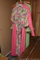 Photo showing the front view of 4 vintage ladies dresses hanging on hangers with one floral pastel dress in front, a bright pink dress behind it, a green dress with ruffle cuffs, and another pink dress visible