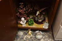 View of inside a cabinet shelf containing a glass bonsai tree, white pope bust, green resin frogs, eagle figurine on a wood base, and a bird music box with a flower-shaped mirror beside it. Below the shelf on a stone floor are a small angel figurine and the bird music box.