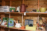 Two wooden shelves inside a shed with various gardening items including garden tools in ceramic pots, bags of fertilizer, a red watering can, a plastic container with drawers, and a boxed garden hose.