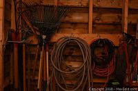 Photo showing garden rakes, hand tools, coiled garden hose, and coiled extension cord hanging on wooden shed wall.