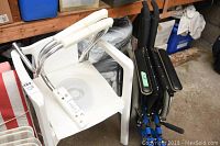 White plastic commode chair with armrests and circular removable seat insert, folded blue wheelchair behind it.