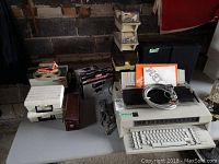 Photo showing IBM Wheelwriter 5 typewriter with cables, manuals, and other boxed items on table in unfinished room