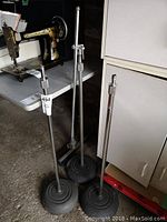Three metal adjustable microphone stands with weighted bases positioned upright near a white cabinet and old sewing machine.