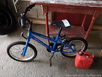 Full view of child's blue Velosport Magic bike and red portable gas can on floor against red workbench.