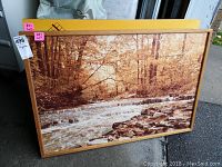 Framed landscape print shown leaning against wall featuring river with flowing water and trees, in oak wood frame.