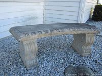 Side angle of concrete bench showing curved seat and carved decorative legs on gravel surface outside beside a house.