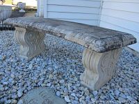 Side angle showing the curved seat with decorative circular pattern along the edge and the carved leg supports with swirling and leaf motifs. Surface shows signs of weather wear and some discoloration.