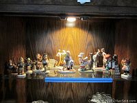 Wide shot of multiple Hummel and Norman Rockwell figurines displayed on a glass shelf under lighting. Various characters and sizes visible.