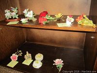 Porcelain floral figurines arranged on two shelves including a large detached floral piece, two lidded containers, and several small floral pieces in white, red, yellow, orange, and pink colors.