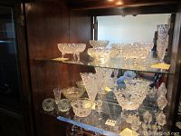Full view of crystal collection on two glass shelves inside a dark wood cabinet, includes bowls, vases, and salt shakers.
