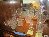 Full set of crystal glassware on wooden shelf including decanter, pitchers, cups, and candle holders.