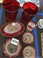 Photo showing two cranberry-colored glass ice buckets with silver rims, cranberry glass covered butter dish, creamer, sugar bowl, and other cut glass pieces on a black table.