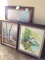 Three framed paintings leaning against a wall including a floral still life, river scene, and countryside cottage