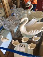 Display of one large white Lenox swan figurine, five smaller Lenox swans, and large Gloria Vanderbilt crystal swan on glass table.