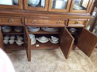 China set displayed in cabinet with doors open showing stacked plates, teacups, and serving pieces including tureen and gravy boat.