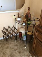 Full view of black metal beverage cart on caster wheels with glass shelves, holding various decanters, ice buckets, small glasses, and a wooden wine rack placed nearby.