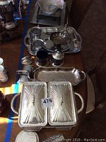 Photo displaying an assortment of silver-plated trays, condiment jars with blue glass inserts, sugar bowl, creamer, and covered serving dishes on a wooden table.