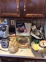 Countertop view of kitchen items including cookie jar, blender, bistro grill box, glass platters, banana-shaped hook and bowl, and assorted kitchenware.