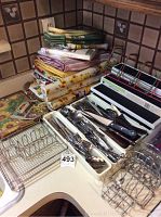 Mixed flatware and kitchen linens stacked on countertop including utensils in silverware tray, dish towels, tablecloths, placemats, and tier curtains.